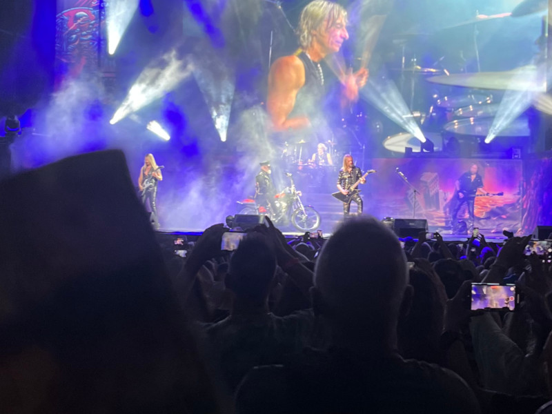 [Image: Rob Halford arrives on stage via motorcycle to join the rest of Judas Priest. The Billy Ripken card is being held up for the camera but is obscurred by the low lighting conditions over the audience.]