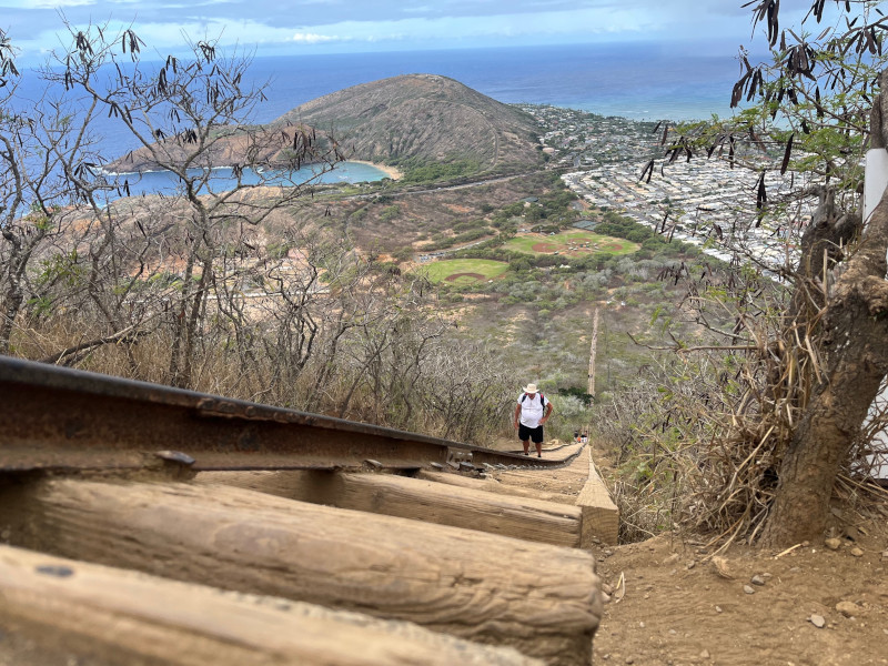 [Image: Photo taken along the Koko Crater tramway looking back towards the trailhead. The headland of Koko Head and the outskirts of Honolulu can be seen as small details in the distance. Baseball diamonds are visible at the bottom of the trail.] 