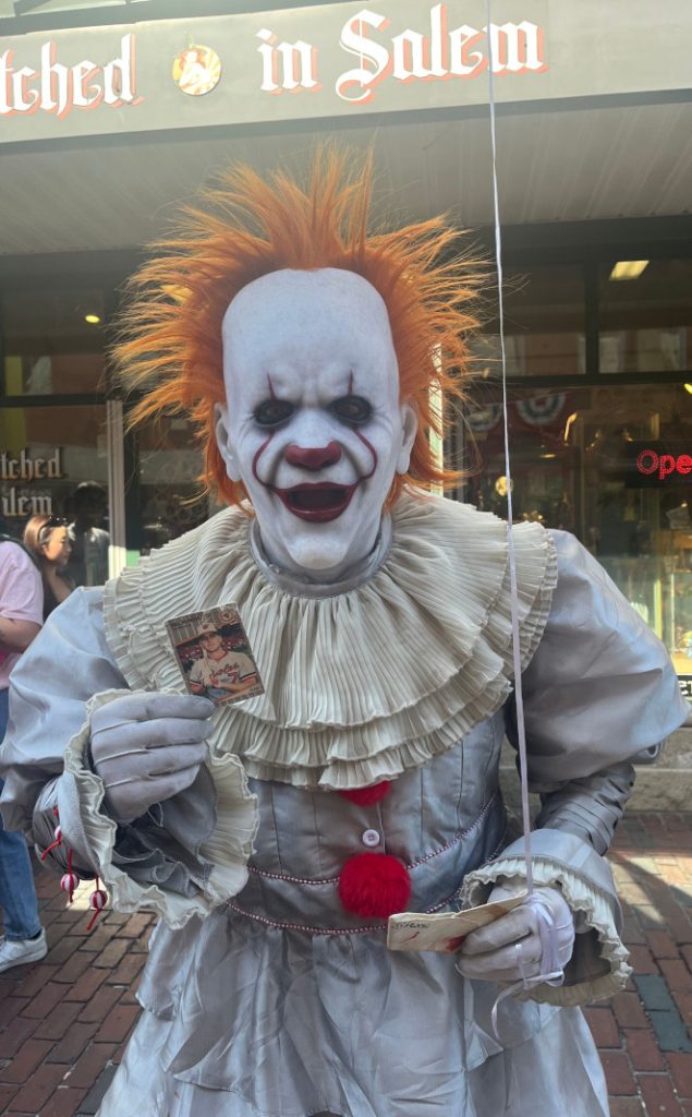 [Image: A cosplayer dressed as Pennywise the Clown holds up the Billy Ripken card for the camera with his right hand. He is holding his red balloon and paper boat in his left hand and standing in front of Bewitched in Salem.]