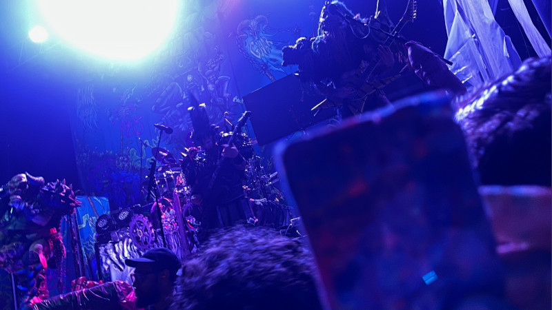 [Image: The Billy Ripken card is held aloft as GWAR performs. The image captures everything in a moment of blue and white stage lighting. Blothar the Berserker is singing on the left, Beefcake the Mighty stands behind him playing bass, and Grodius Maximus plays guitar directly in front of the person taking the picture.]