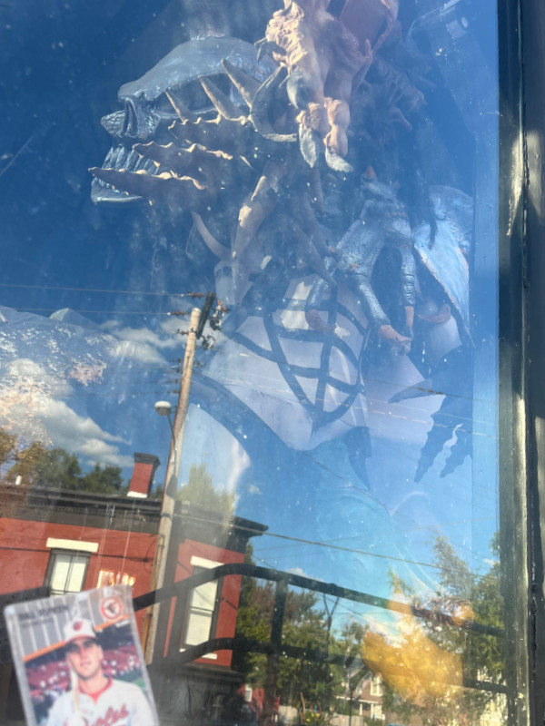 [Image: The Billy Ripken card is propped against a second story window at GwarBar. On the other side of the glass is a mannequin wearing the stage armor of Oderus Urungus, complete with Cuttlefish of Cthulhu. The image has a large amount of reflective glare.]