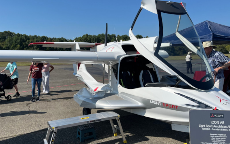 [Image: A parked Icon A5. The canopy is open and the left wing is folded back in the parked position. The plane's single rear propeller is visible. An informational sign about the aircraft is present.]