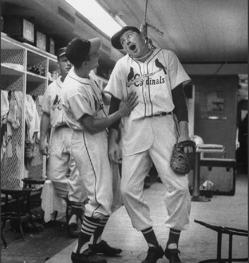 [Image: Frank Smith is seen standing on his tip toes with a noose from the ceiling around his neck, grinning through mock agony at teammate Stan Musial who is checking on his safety. Another Cardinal player is approaching the scene from behind with a genuine look of concern. Image captured by Francis Miller, used under personal non-commercial terms.]